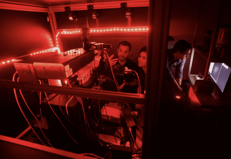 3 scientists working in a small lab space that is illuminated by red lights