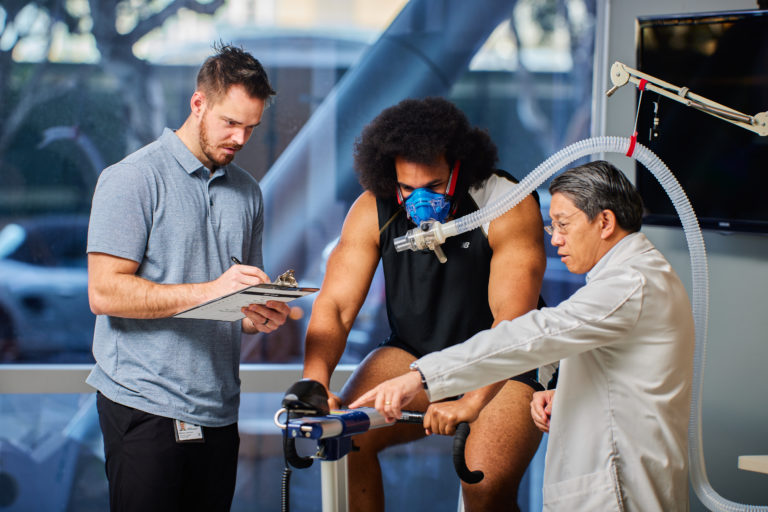 Human Performance Lab image: a strong person on a stationary bike has an air mask over their nose and mouth. 2 people point out and record data