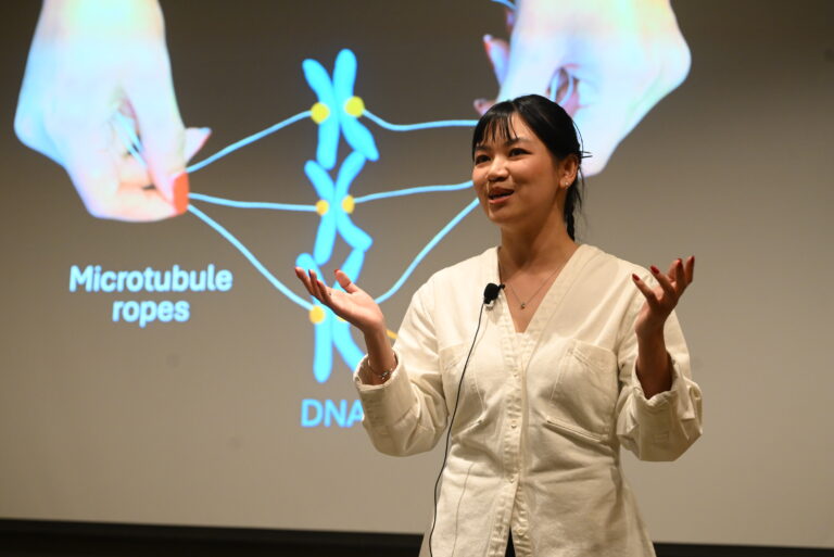 A young female scientist stands with her hands in an outward gesture in front of her slide that reads Microtubule ropes and DNA