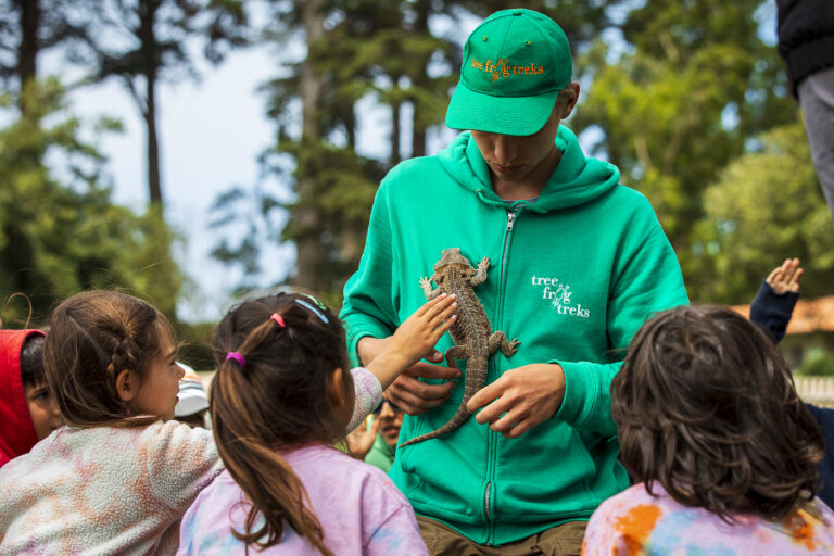 An adult in a green sweatshirt and matching hat holds a bearded dragon lizard while kids reach out to feel its skin