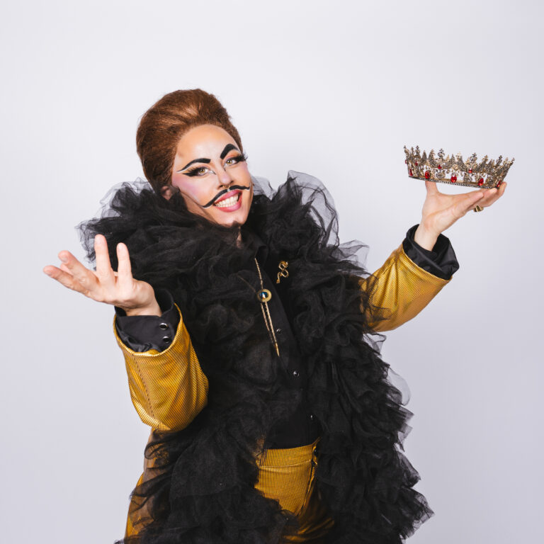Drag King Misterrr posing in a yellow suit with a giant black scarf, holding a crown with red jewels. Thick mustache and eyebrown makeup.