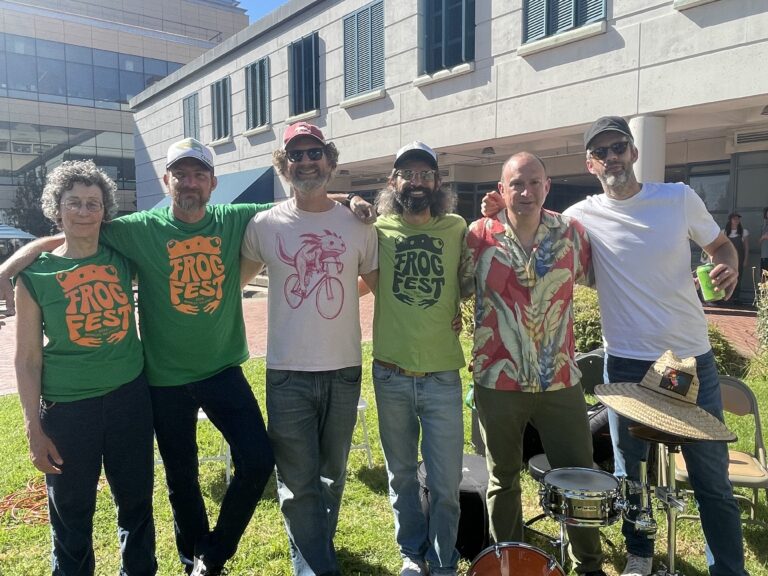 6 bandmates smiling in the sun on a grassy lawn. Drum kit is in front of them.