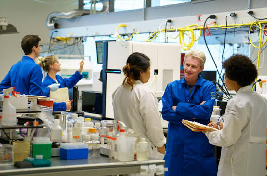 Joe DeRisi in the lab working with 4 scientists dressed in white and blue lab coats